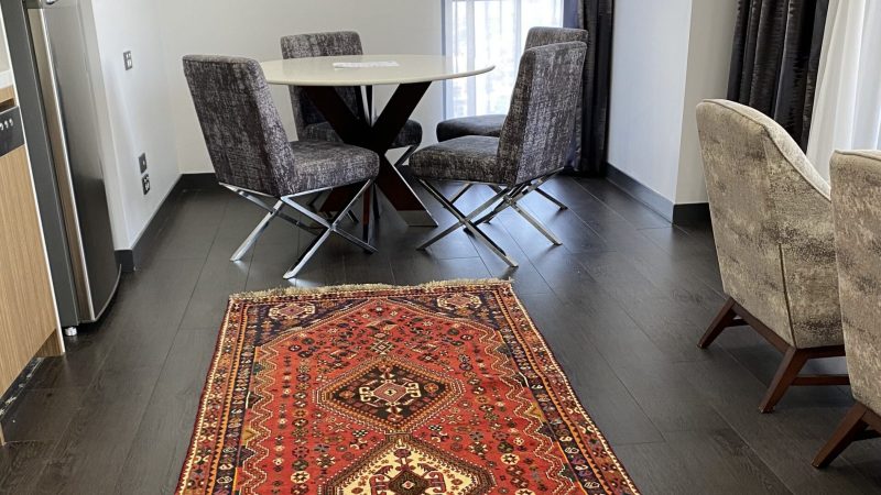 Image description: A photograph of a Persian rug placed on a hardwood floor. The rug is rectangular and features intricate patterns and designs in earthy colors such as beige, brown, and navy blue. The patterns consist of floral and geometric shapes arranged symmetrically. The rug has a medium pile and is slightly folded on one side, adding depth and texture to the image. The warm lighting in the room highlights the rug's details and creates shadows. This image is intended to illustrate how a Persian rug can add beauty and style to a room's decor.