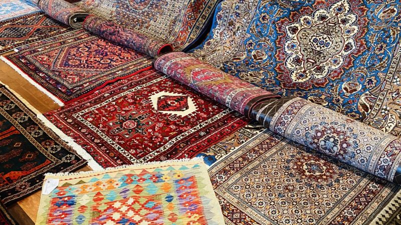 A close-up of a traditional Persian rug with an intricate design of floral and geometric patterns in warm colors of red, orange, and gold, on a neutral background.