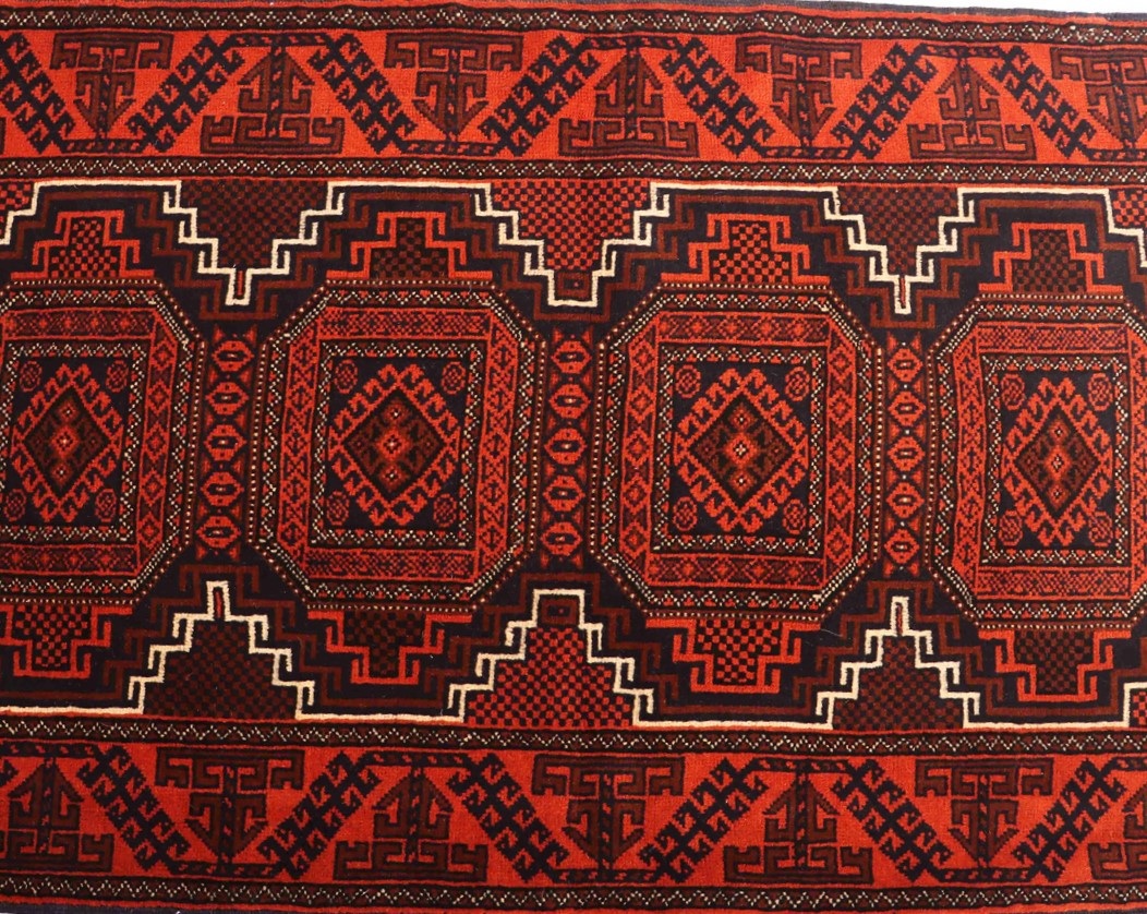 A close-up image of a handmade Balouchi rug featuring a symmetrical pattern with a vibrant color palette of red, blue, and cream. The intricate design includes diamond-shaped motifs and geometric patterns. The rug is placed on a hardwood floor with natural lighting casting shadows over its texture.