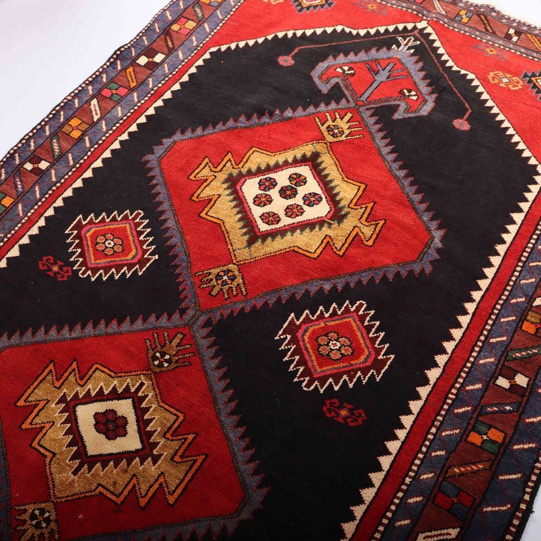 A close-up photo of a Persian rug with intricate geometric patterns and bright colors, lying on a wooden floor. The rug features a central diamond-shaped medallion in red, surrounded by smaller diamond-shaped motifs in blue, green, and beige. The edges of the rug are adorned with a series of thin borders in various colors and patterns.